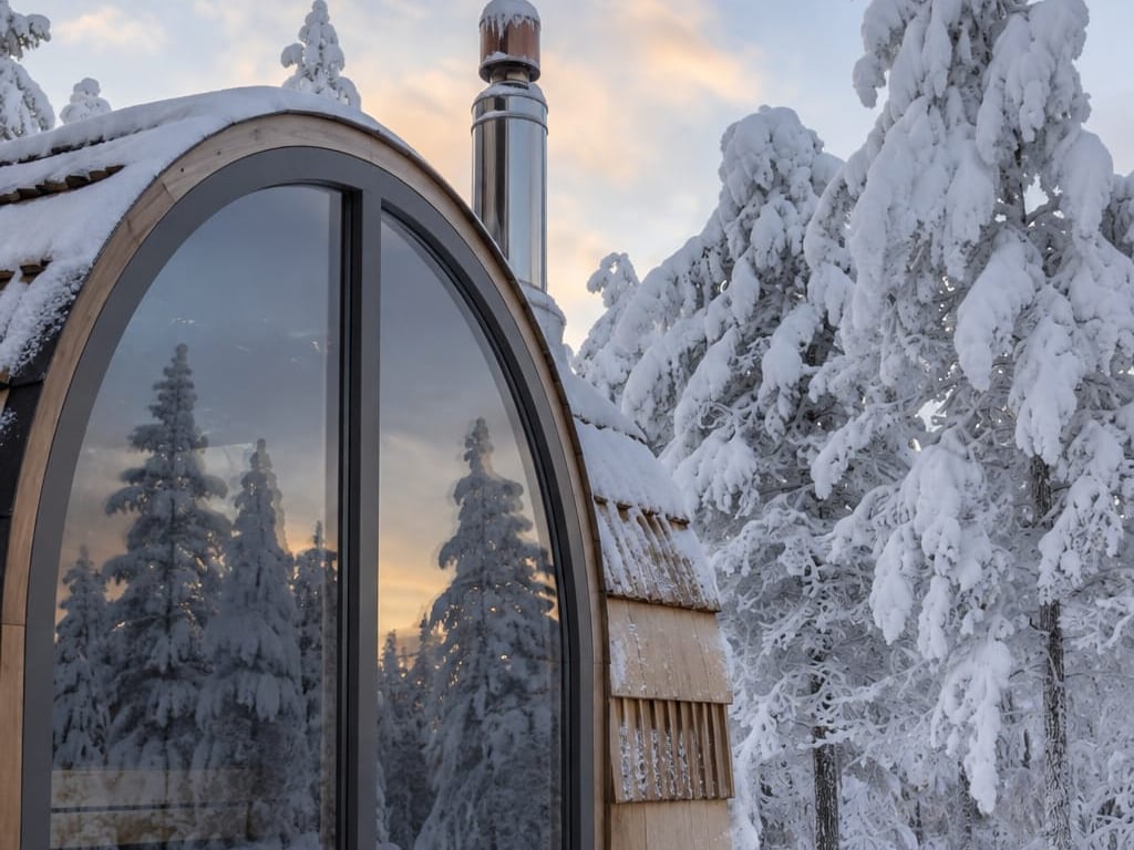 Snow-covered sauna with an arched window reflecting the winter forest.
