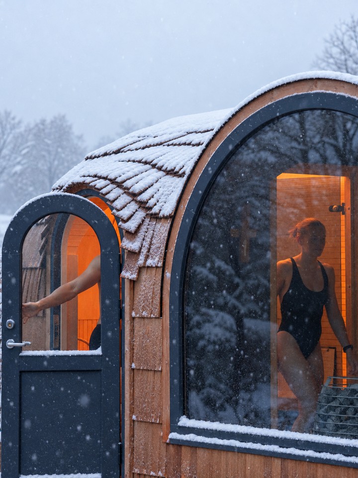 Winter visualization of an Iglucraft sauna with curved timber shingles, a panoramic window and a warm interior.